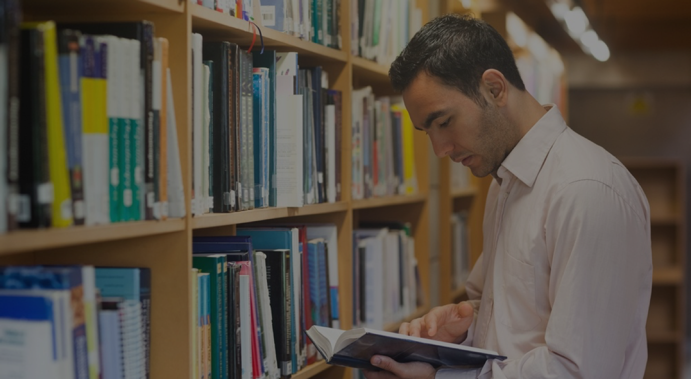 Dark_Intellectual man reading a book standing in library in front of bookshelves.png
